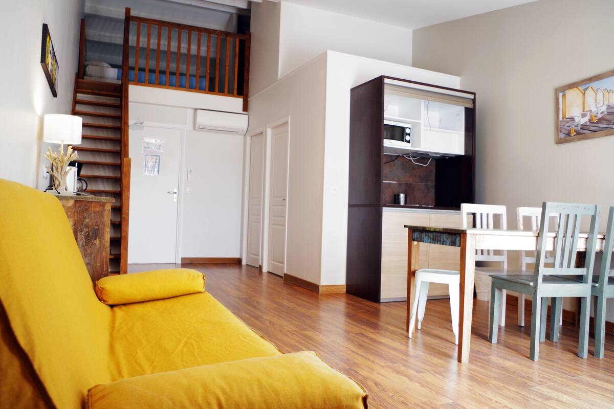 A living room with a yellow sofa, a dining area with white chairs, a kitchen with dark cabinets, and a wooden staircase.
