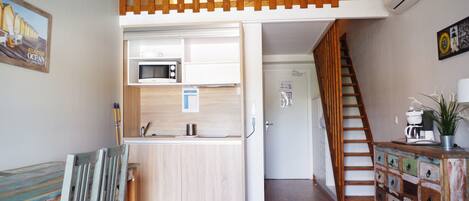 A compact kitchen with a microwave, wooden cabinets, and a small dining area with a chair.