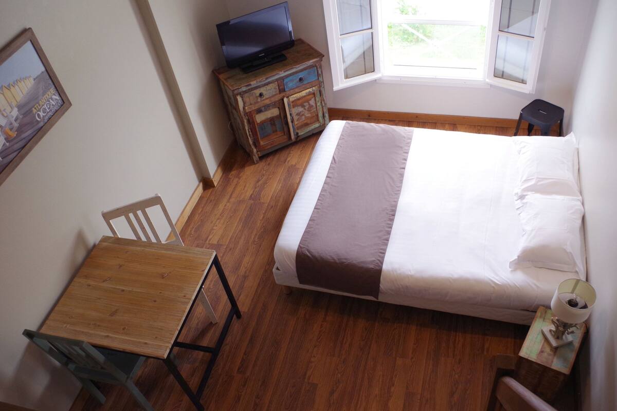 A bedroom with a wooden floor, a bed with white and brown bedding, a wooden nightstand with a lamp, a small wooden table with a chair, and a television on a wooden dresser.
