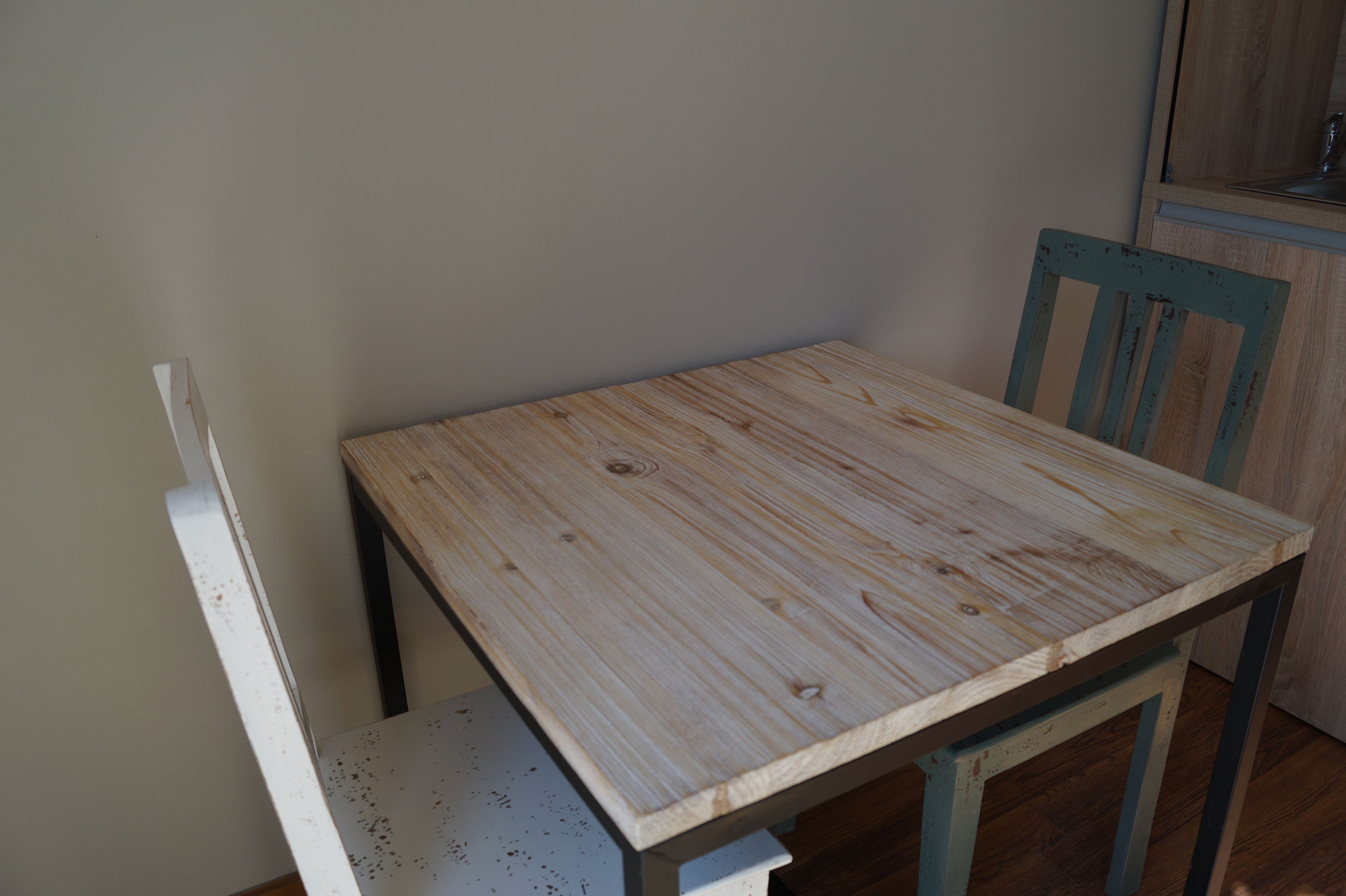 Una mesa de madera pequeña con una silla, un banco blanco y un armario de madera en una habitación de paredes lisas.