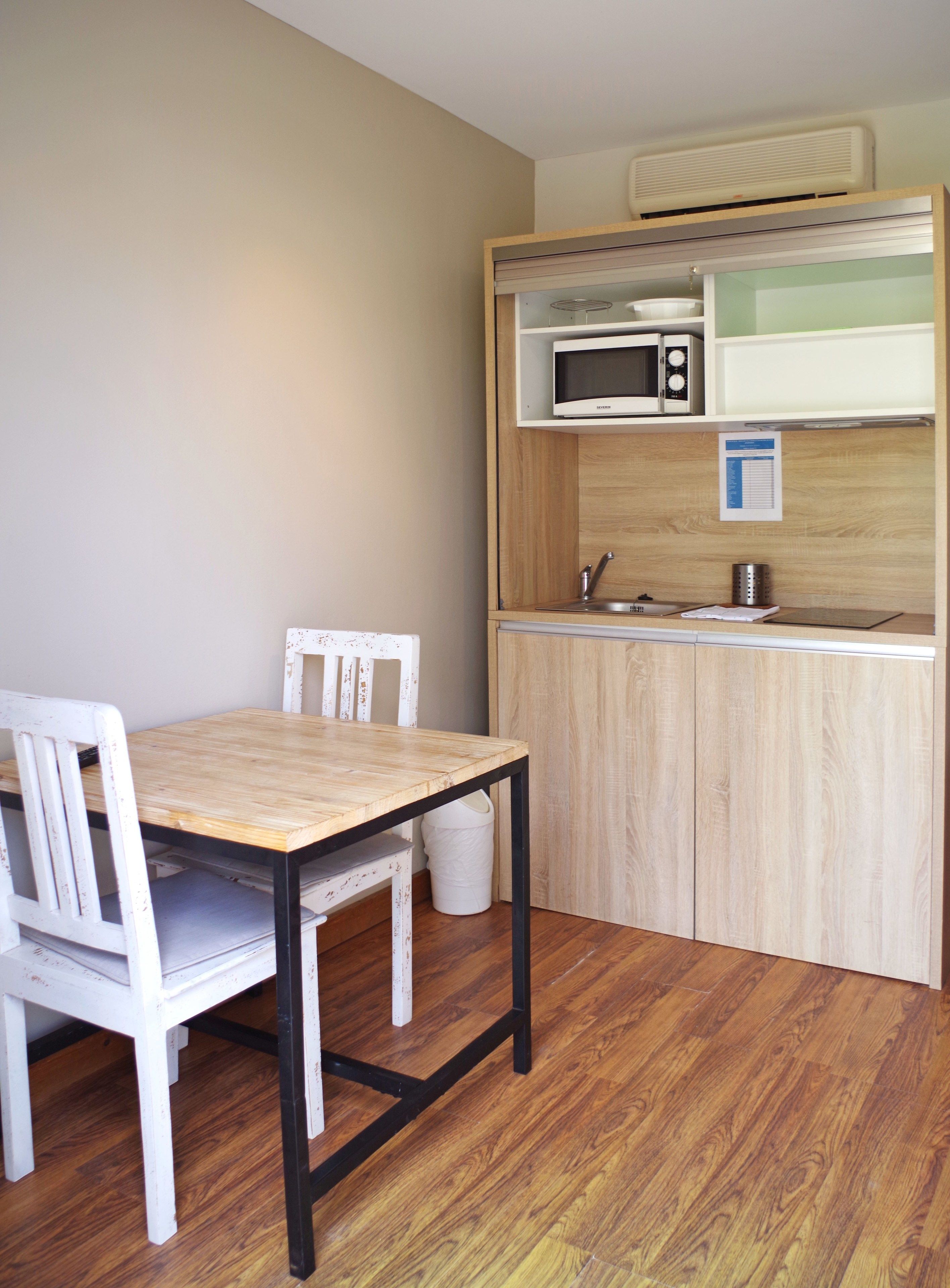 Een compacte keuken met een magnetron, spoelbak en een klein kastje. Er is een houten tafel met twee stoelen en een afvalbak.