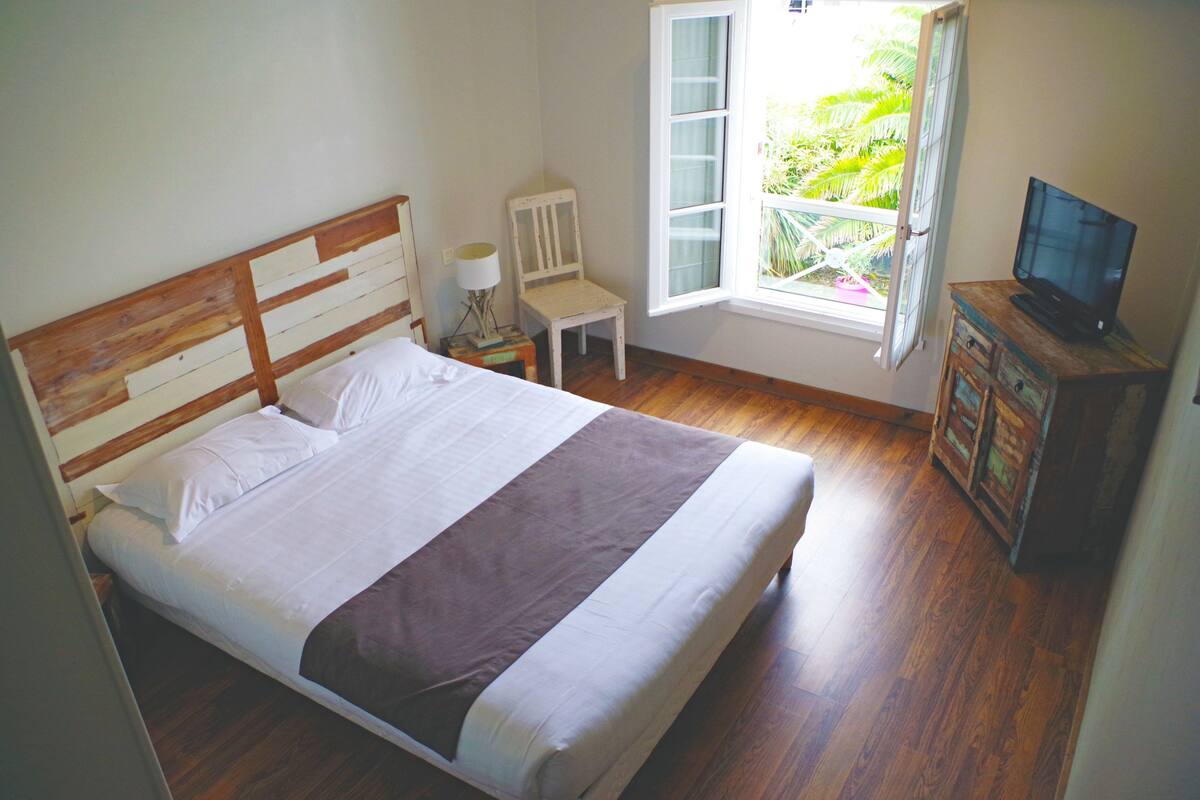 A neatly made bed with a wooden headboard, a small bedside table with a lamp, a chair by the window, and a television on a wooden cabinet.