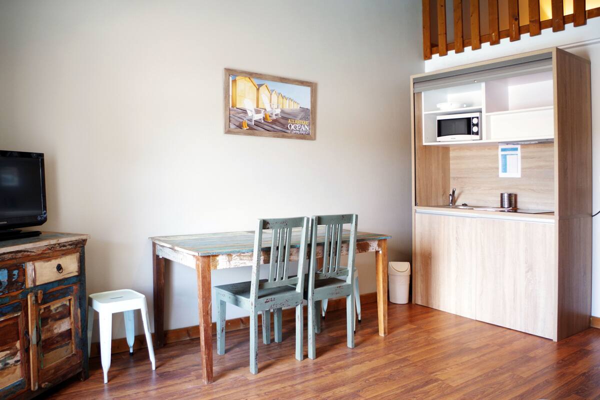 A compact living space with a wooden dining table, two chairs, a small white stool, a television on a wooden cabinet, and a kitchenette with a microwave.