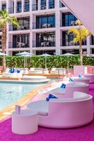 Zona junto a la piscina con sillones blancos, piscina y un edificio con balcones al fondo.