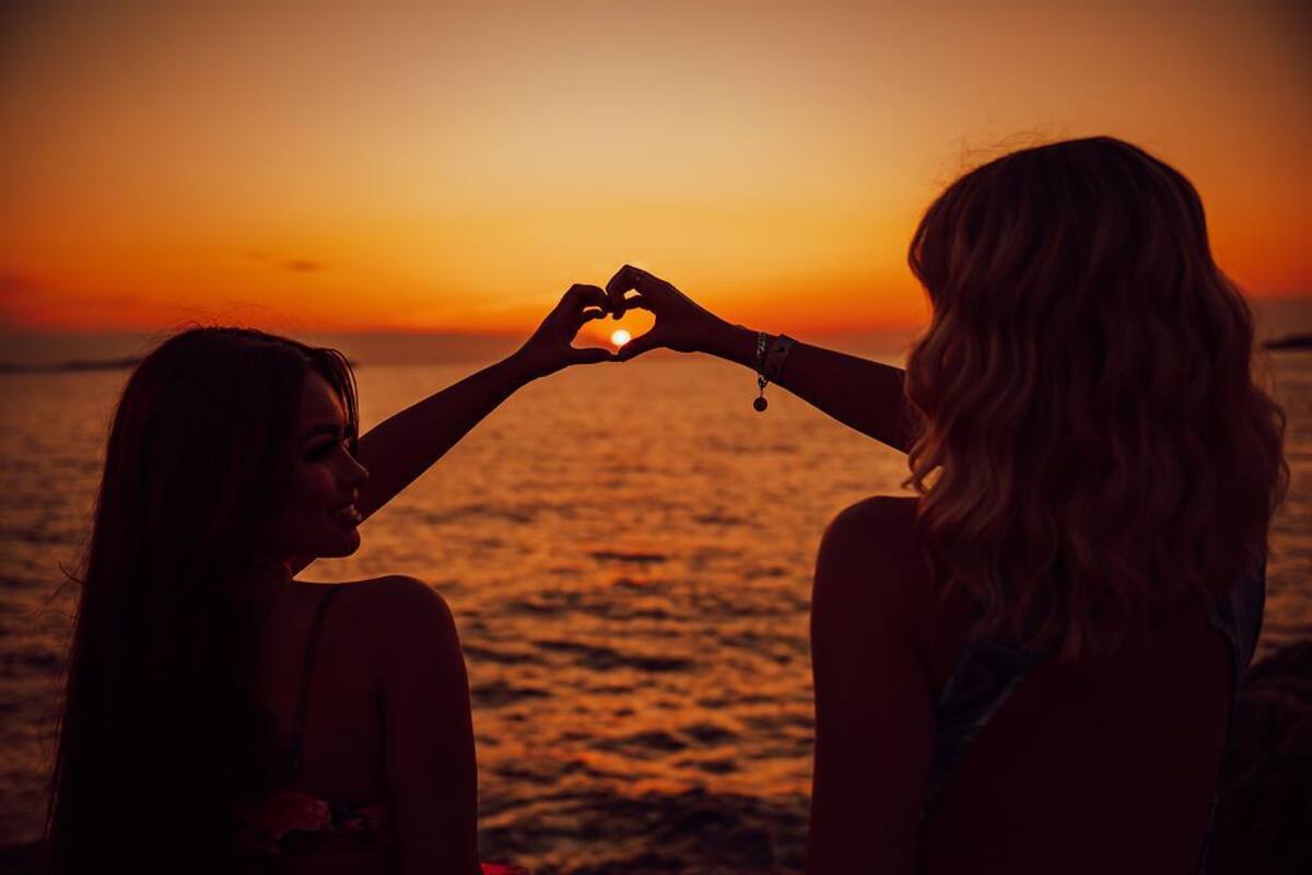 Two individuals making a heart shape with their hands against a sunset backdrop.