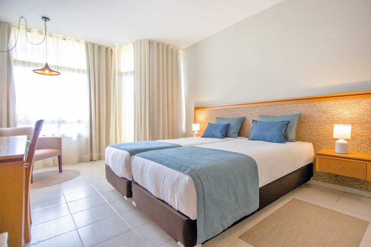Quarto de hotel com duas camas, uma escrivaninha e uma cadeira.