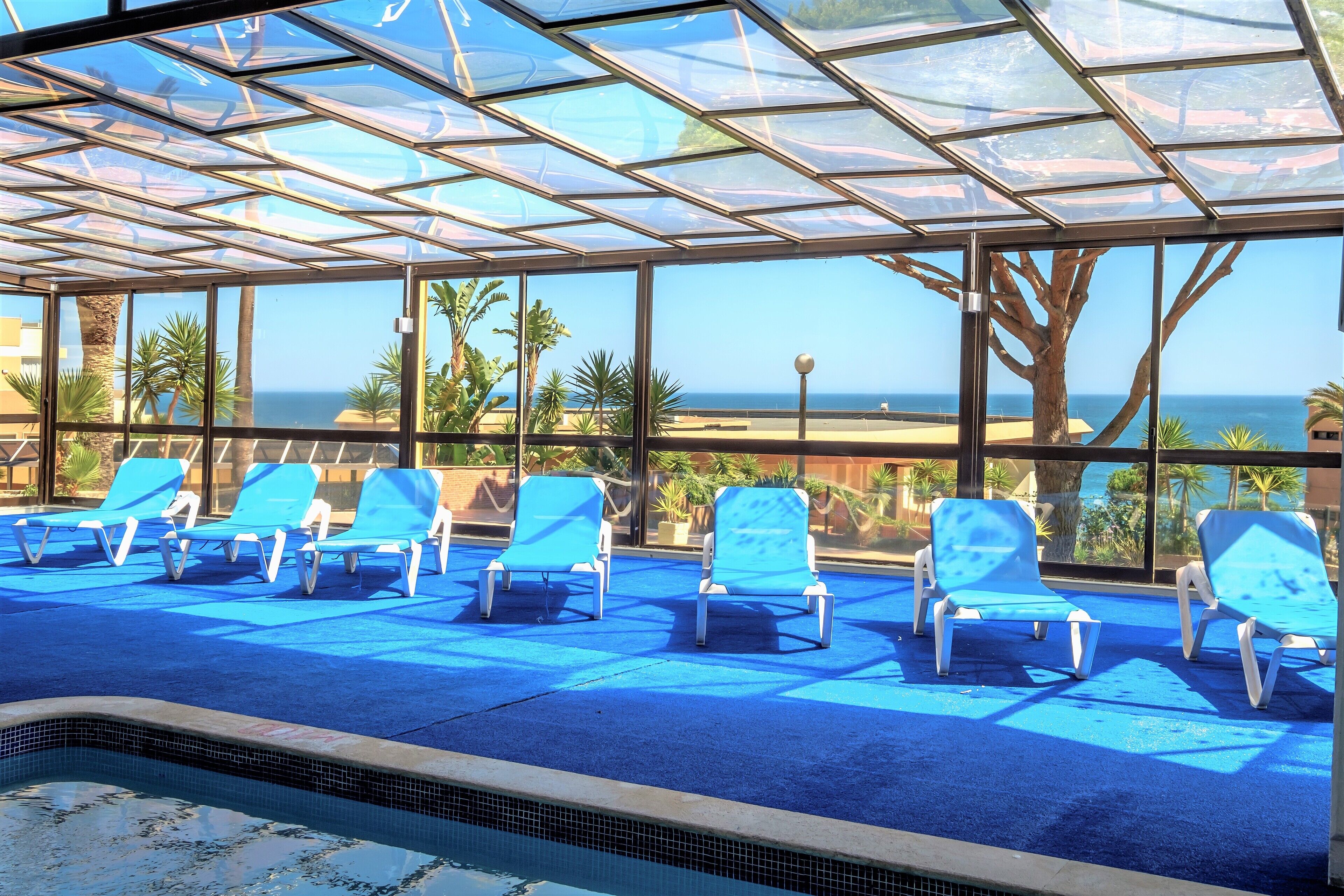 Zona de piscina con sillas reclinables azules y techo de vidrio con vista al océano.