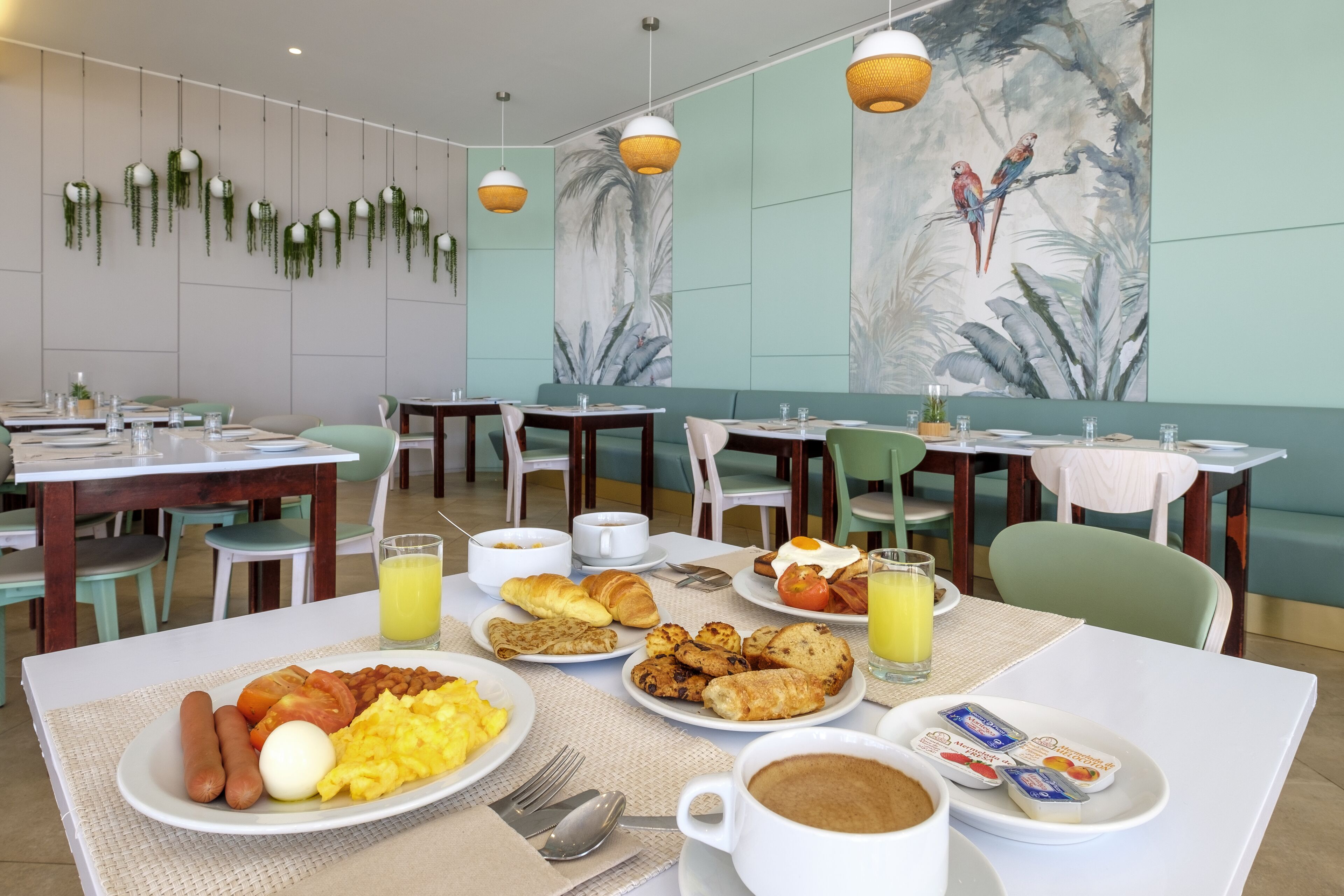 Un buffet de petit-déjeuner proposant divers plats, notamment des œufs, des saucisses et des viennoiseries, dans un espace de restauration agrémenté de chaises et de tables vertes.