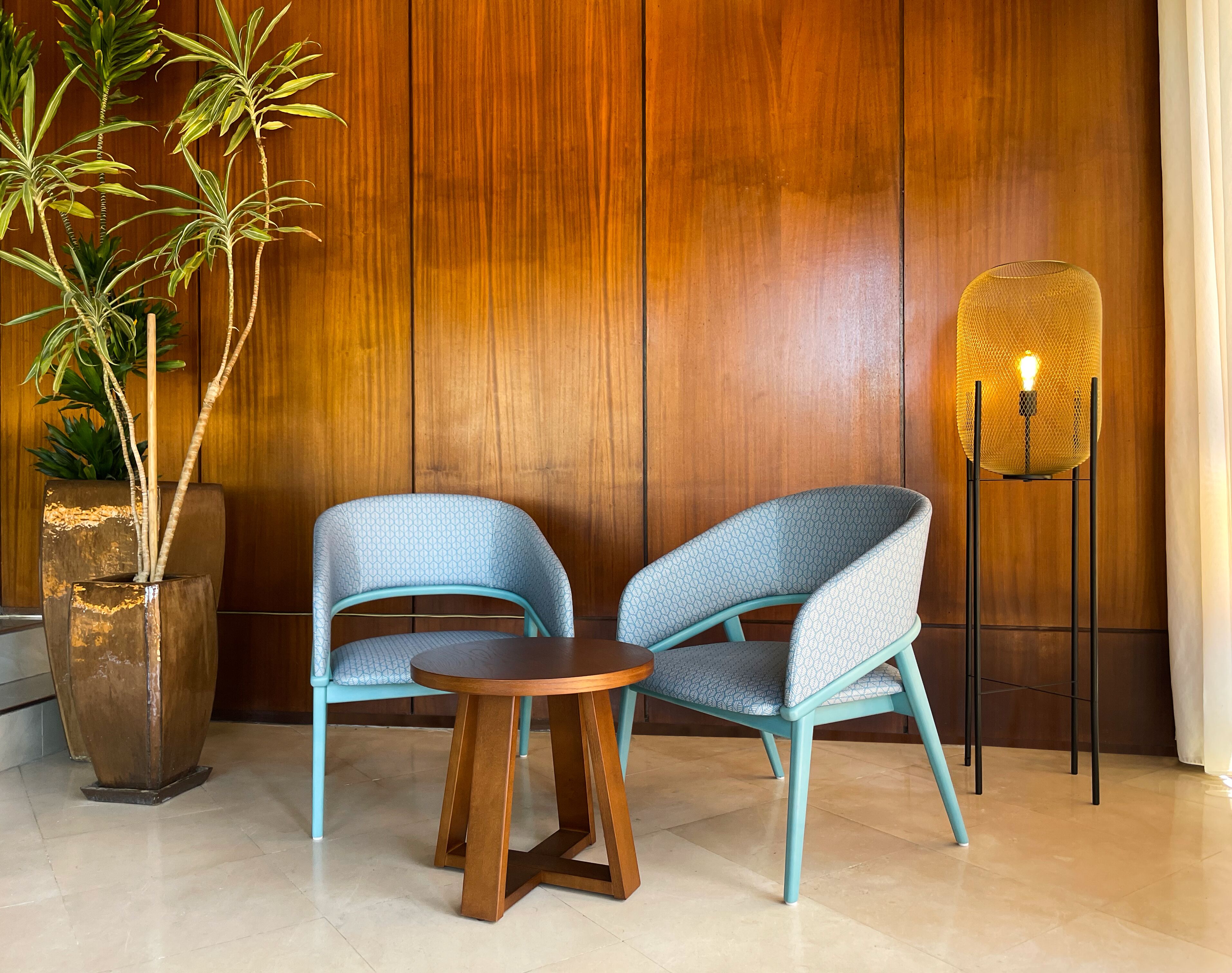 Una habitación con dos sillas azules con diseño, una mesa de madera, una lámpara de pie alta y una planta grande en maceta frente a una pared de madera.