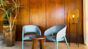 Una habitación con dos sillas azules con diseño, una mesa de madera, una lámpara de pie alta y una planta grande en maceta frente a una pared de madera.