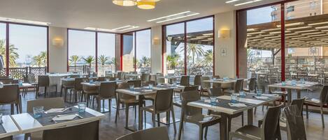 A spacious dining area with multiple tables and chairs, large windows offering views of palm trees and buildings, and modern lighting fixtures.