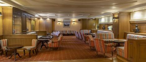 A spacious hotel lobby with wooden paneling, striped upholstered seating, and a central area with tables and chairs.