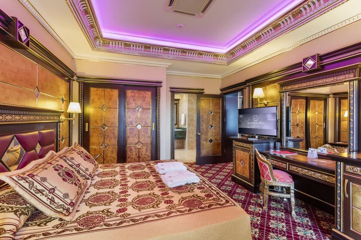 A luxurious hotel room with a large bed, a desk with a television, and a bathroom visible through an open door.