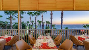 An outdoor dining area with a table set for two, overlooking a beach with palm trees and the ocean at sunset.