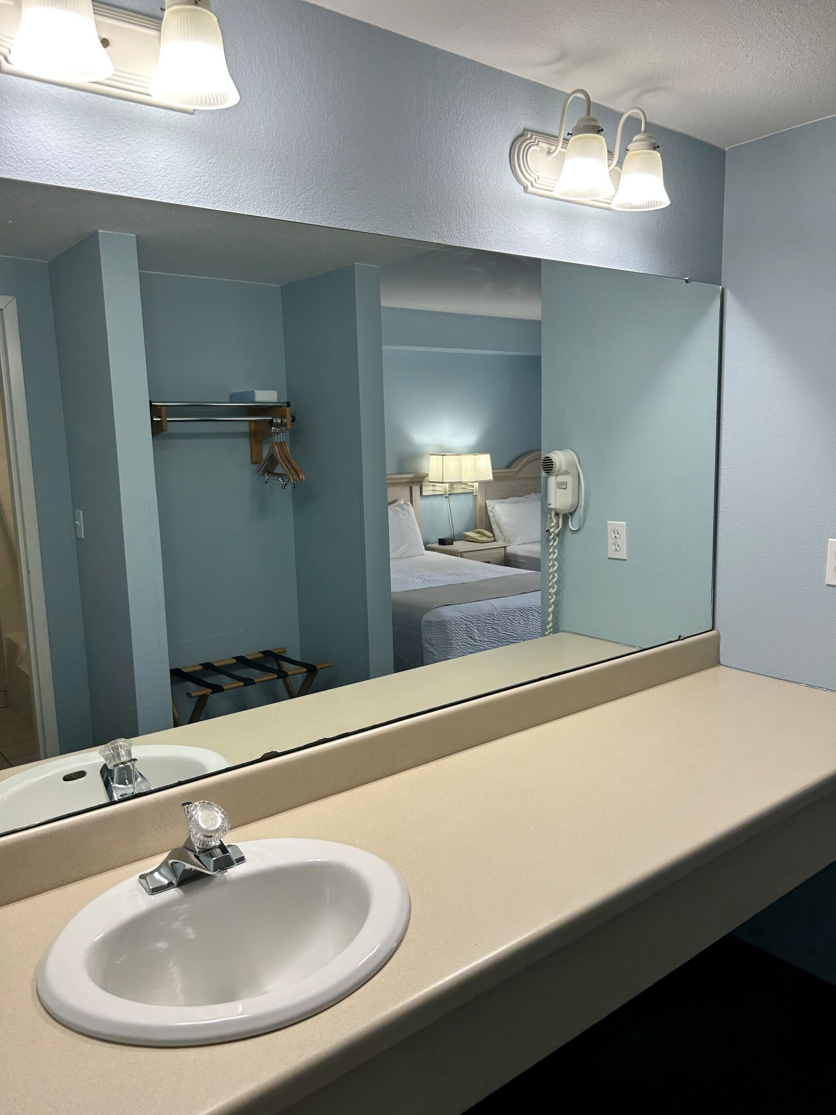 A bathroom with a large mirror, two sinks, a towel rack, and a bed visible in the reflection.