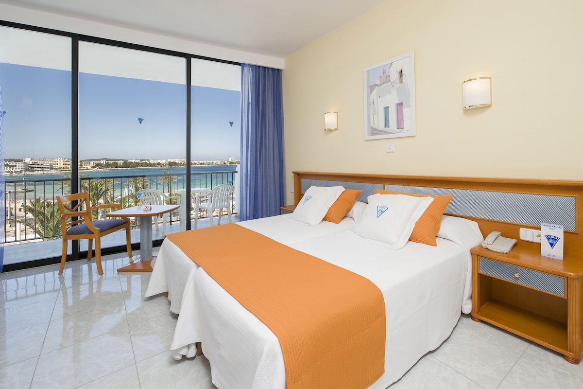 Habitación de hotel con una cama grande, una mesita de noche, una silla, una mesa y vistas al mar.