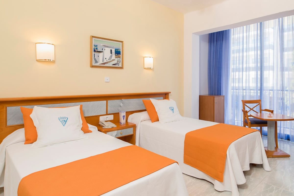 Habitación de hotel con dos camas, un cabecero de madera, una mesita pequeña, una silla y un ventanal grande con cortinas.