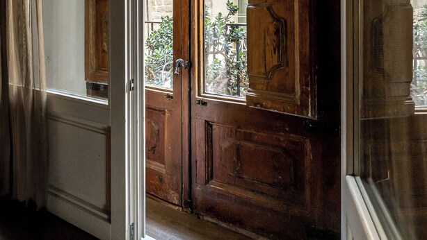 Puerta de madera con un panel de vidrio, que da a un espacio exterior con pared de piedra y un balcón.