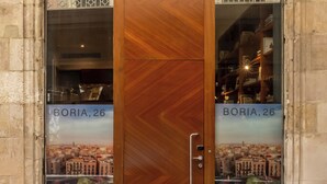 Una puerta de madera con manija y paneles de vidrio, que presenta un cartel con un paisaje urbano y el texto "BORIA, 26".
