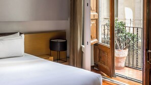 Un dormitorio con cama, mesita de noche, lámpara y vistas a un balcón exterior con una planta en maceta.
