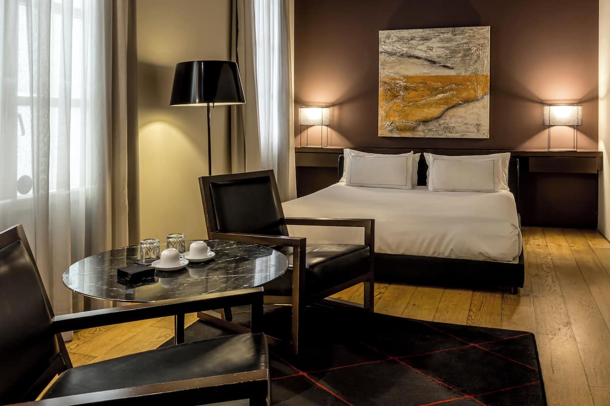Ein Hotelzimmer mit einem Bett, zwei Nachttischlampen, einem Bild an der Wand, einem Tisch mit Stühlen und einem Fenster mit Vorhängen.