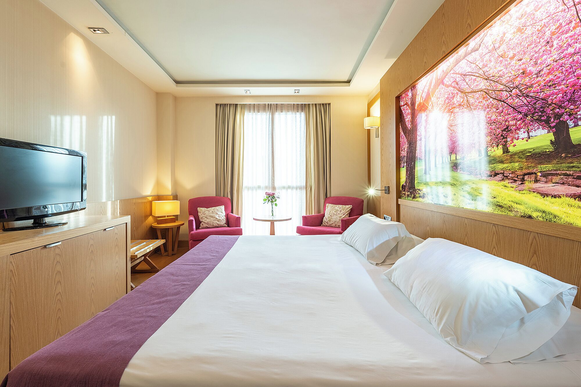 Une chambre d’hôtel avec un grand lit, un téléviseur posé sur une commode en bois, deux fauteuils, une petite table avec une lampe et une grande fenêtre avec des rideaux.