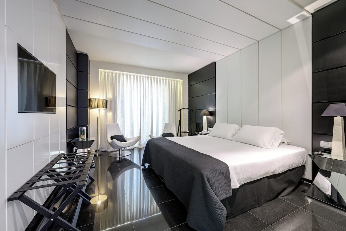 Quarto de hotel moderno com uma cama grande, uma escrivaninha com cadeira, um abajur e uma janela ampla com cortinas.