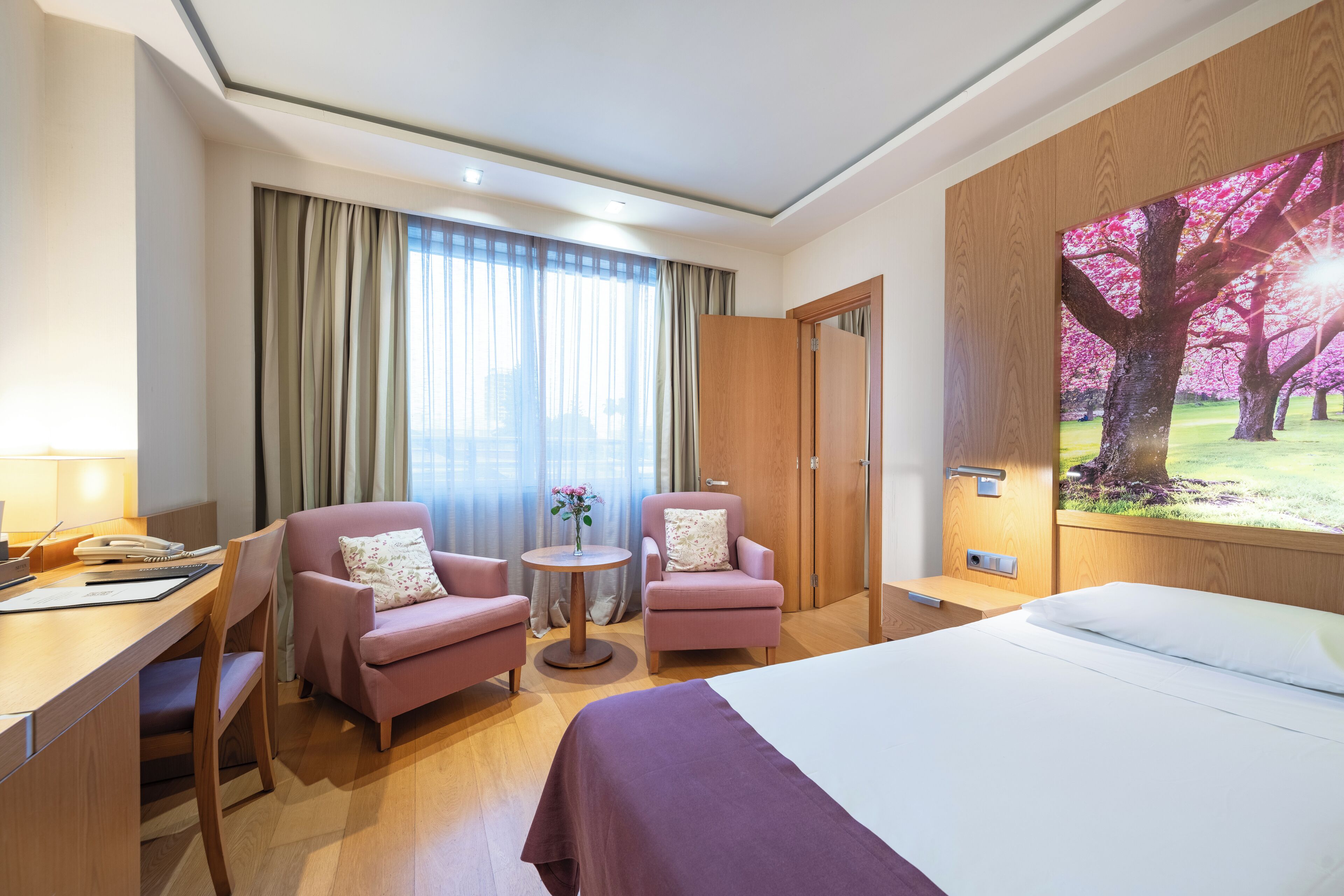 Ein Hotelzimmer mit einem Bett, zwei Sesseln, einem Schreibtisch mit Telefon und einem kleinen Tisch mit Blumen.