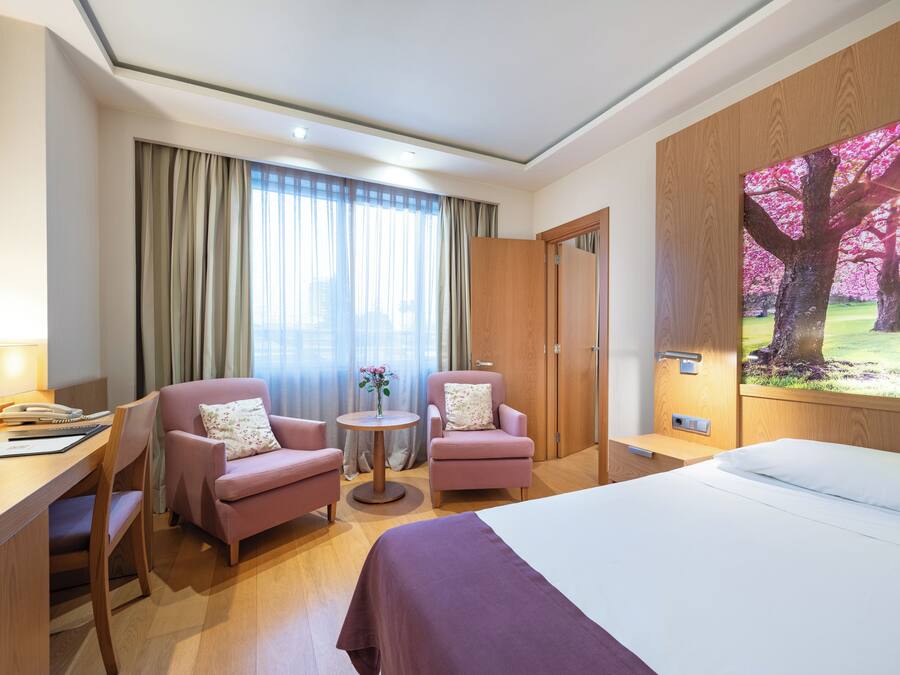 Habitación de hotel con una cama, dos sillones, un escritorio con teléfono y una pequeña mesa con un florero.