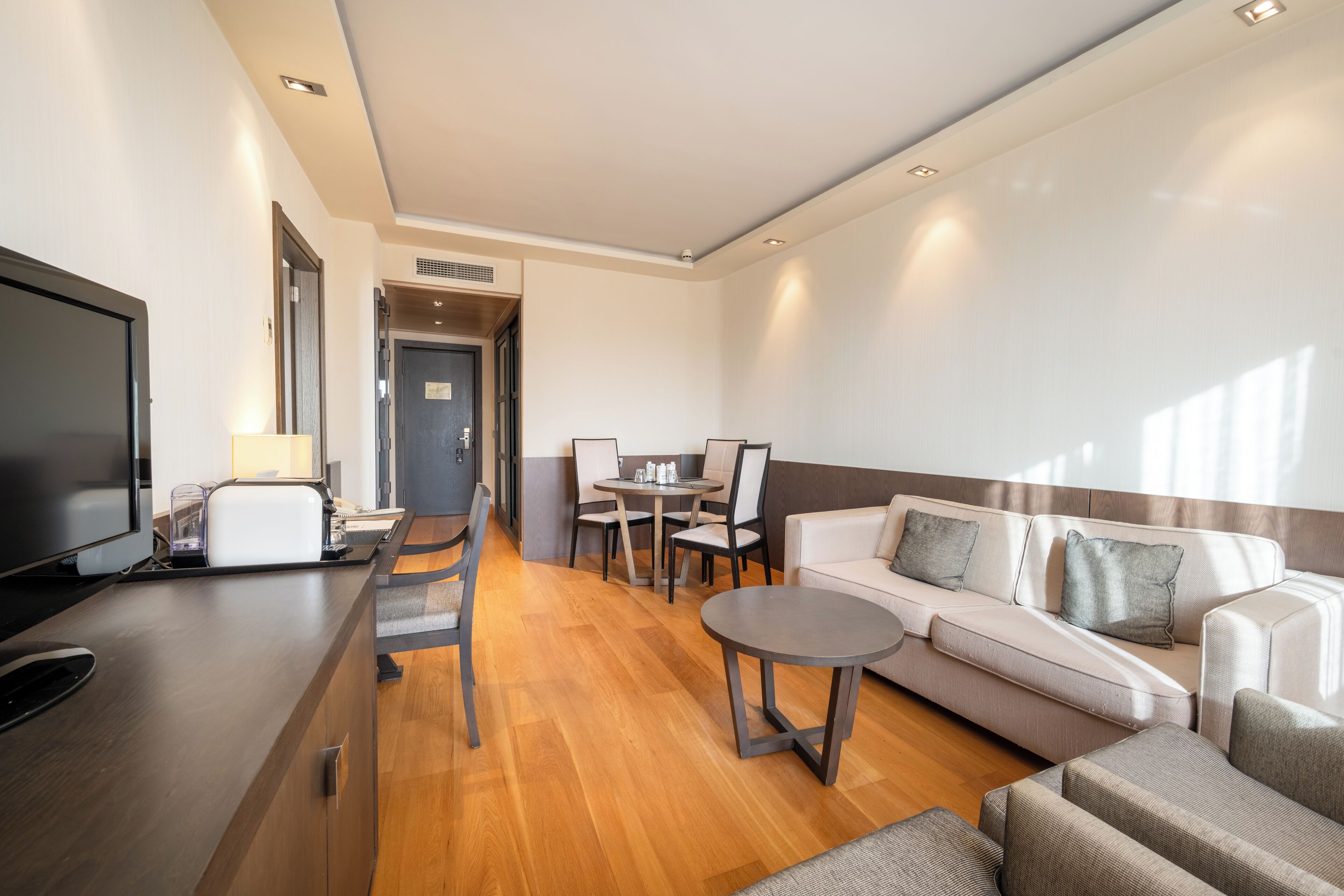 A modern hotel room with a wooden floor, a sofa, a TV, a dining table with chairs, and a small round table.