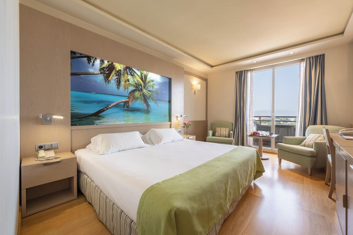 Quarto de hotel com uma cama grande, uma escrivaninha, uma cadeira, uma mesinha e uma varanda com cortinas.