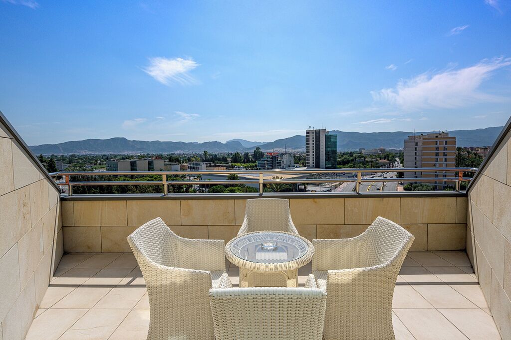 Varanda com cadeiras de vime e mesa de vidro, com vista para uma paisagem urbana e montanhas ao longe.