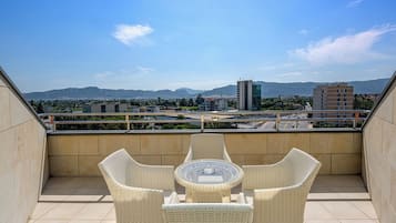 Un balcón con sillas de mimbre y una mesa de vidrio, con vistas a un paisaje urbano y montañas al fondo.