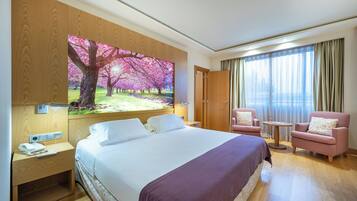A hotel room with a large bed, a desk with a telephone, a chair, a small table, and a large window with curtains.