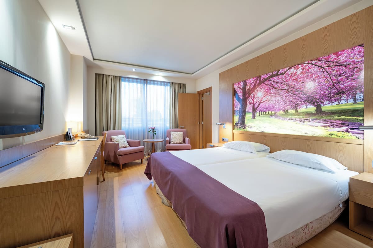 Quarto de hotel com uma cama grande, uma escrivaninha, uma televisão, uma cadeira e um quadro de flores de cerejeira.