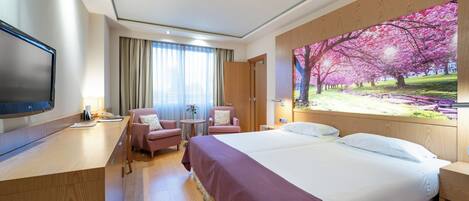 A hotel room with a large bed, a desk, a TV, a chair, and a painting of cherry blossoms.
