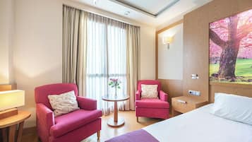A hotel room with two pink armchairs, a round table with a vase, a bed with white linens, and a painting of a tree.