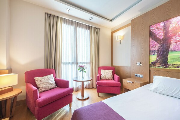 Habitación de hotel con dos sillones rosados, una mesa redonda con un florero, una cama con ropa blanca y un cuadro de un árbol.