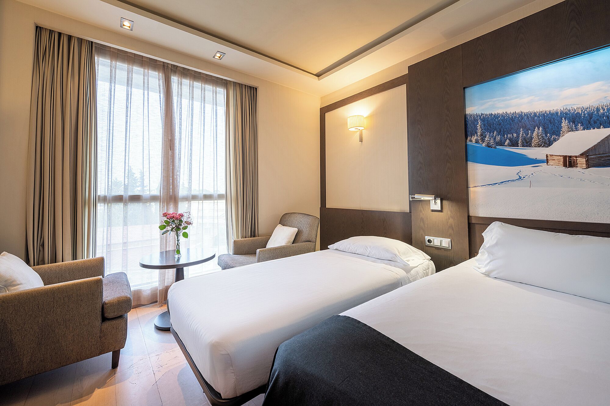 Habitación de hotel con dos camas, una silla, una mesita con un florero y un ventanal con cortinas.