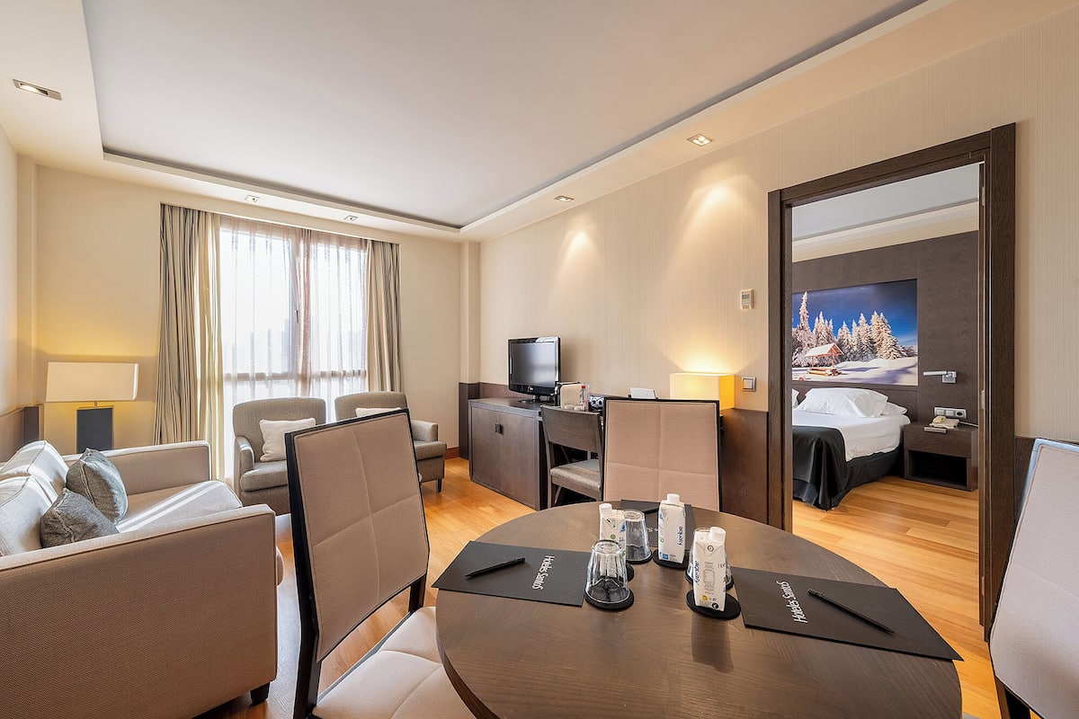 Quarto de hotel com área de refeições, televisão, cama e pintura de uma paisagem nevada.