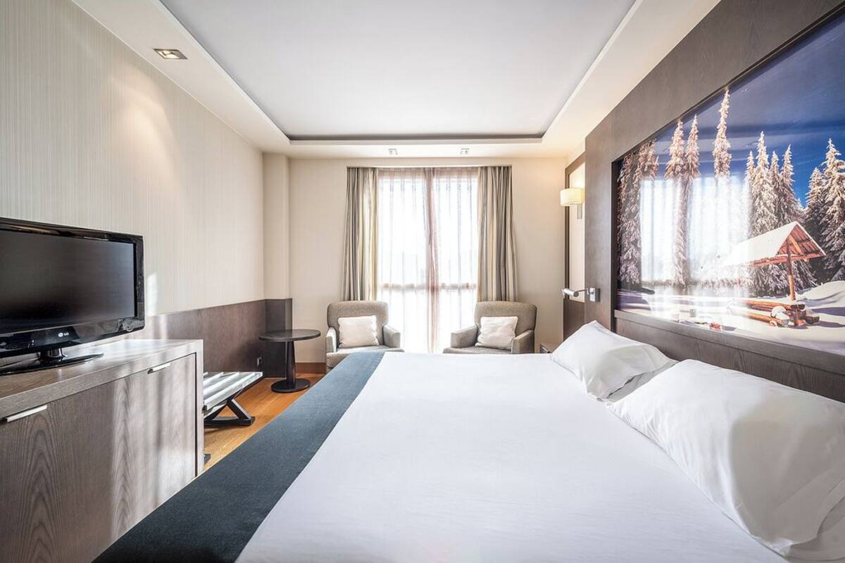 Quarto de hotel com cama, televisão, duas cadeiras, escrivaninha e pintura de uma paisagem nevada.