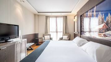 Habitación de hotel con cama, televisor, dos sillones, escritorio y un cuadro de un paisaje nevado.
