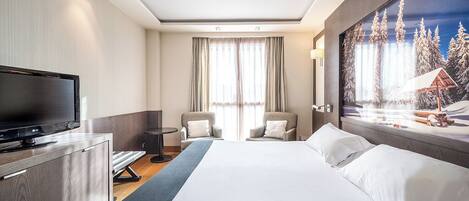 A hotel room with a bed, a TV, two chairs, a desk, and a painting of a snowy landscape.