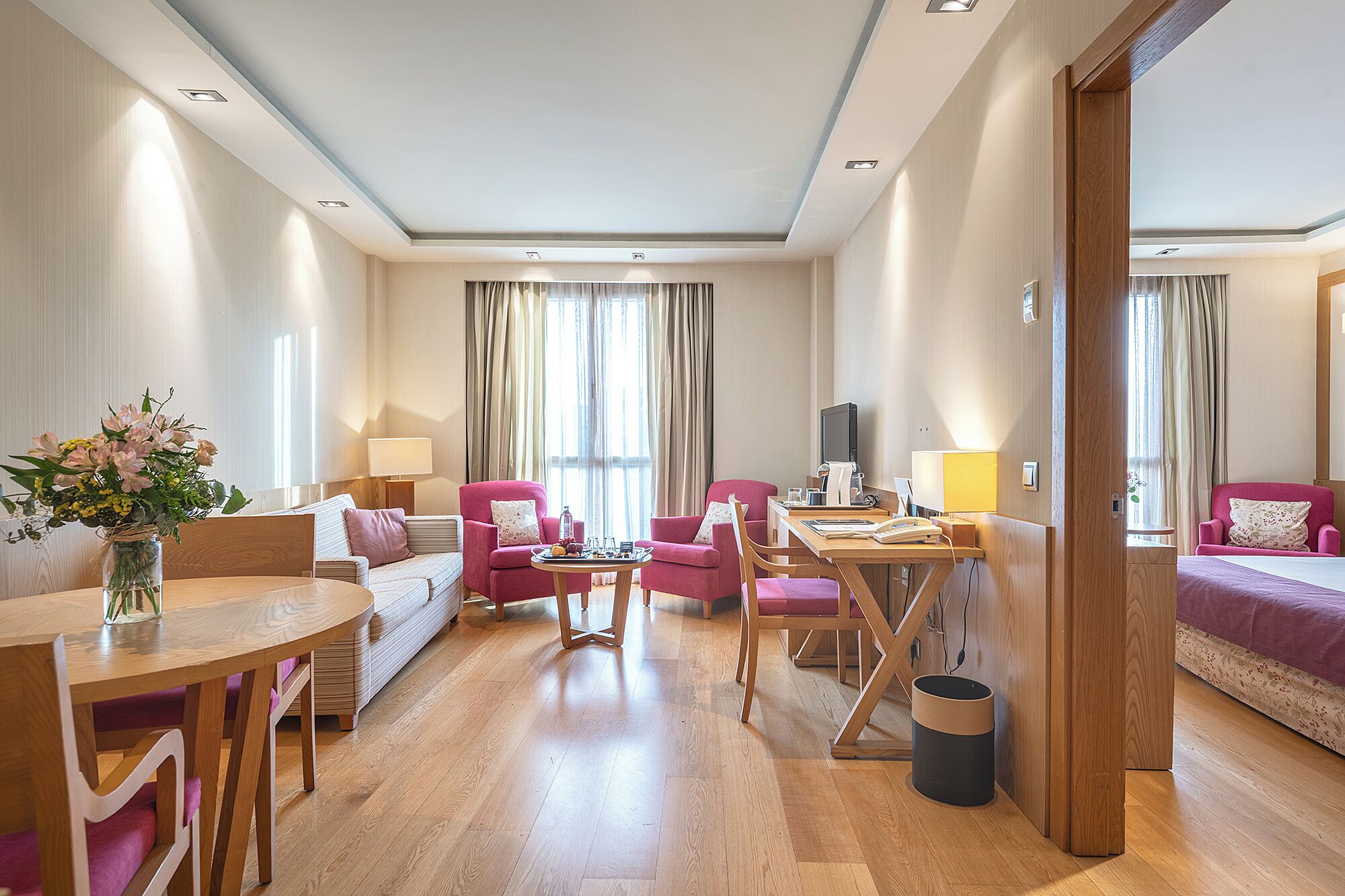 A hotel room with a wooden floor, a dining table, two pink armchairs, a sofa, a TV, and a vase of flowers.