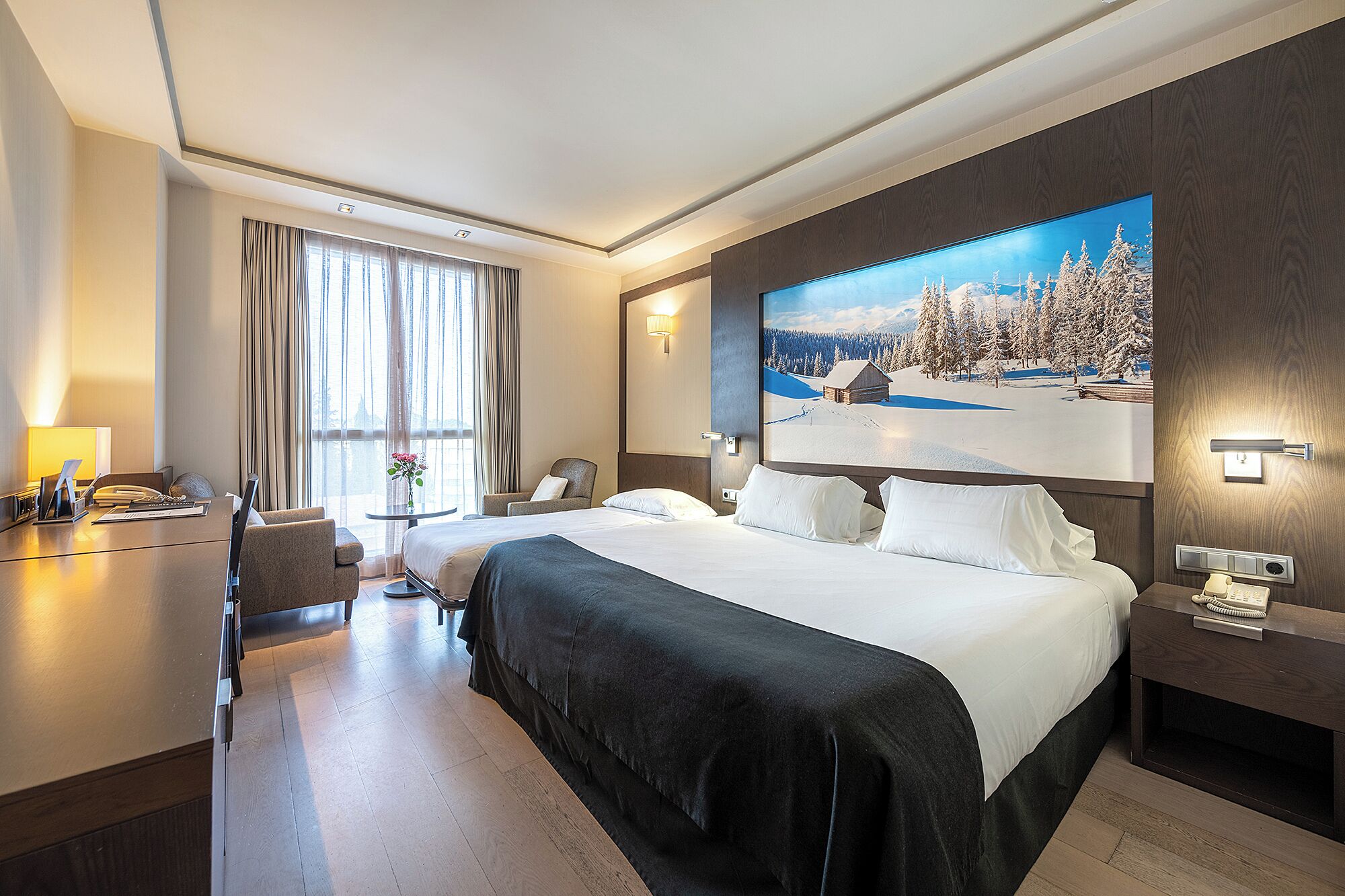 Habitación de hotel con una cama grande, un escritorio con una lámpara, una silla, una mesita con un jarrón y un cuadro de un paisaje nevado en la pared.