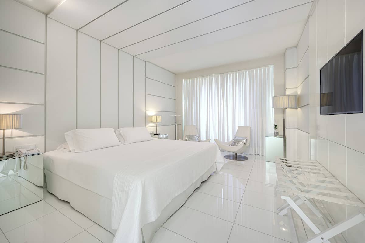 Quarto de hotel moderno com uma cama grande, mesas de cabeceira, uma cadeira e televisão fixada na parede.