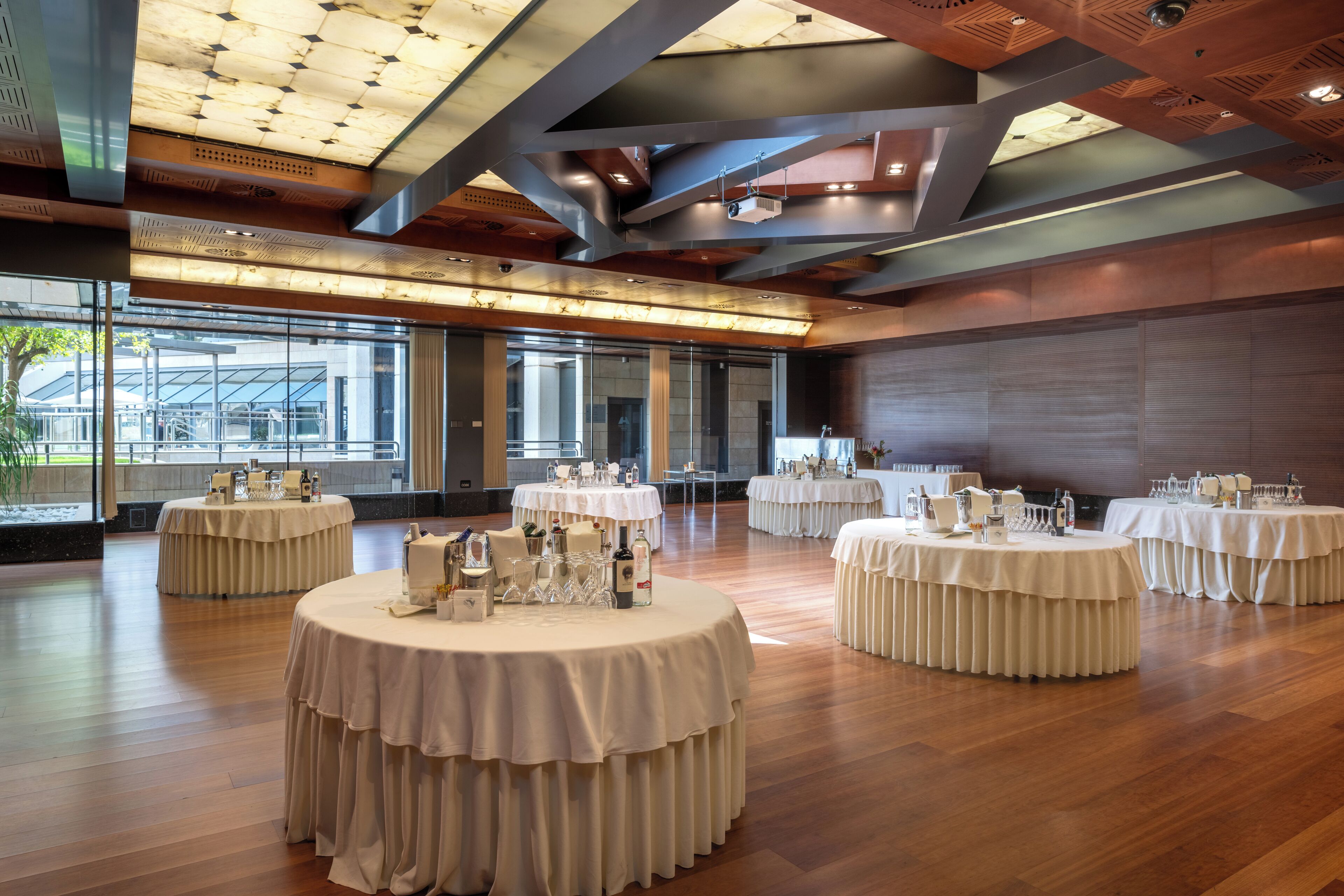 A spacious hall with multiple round tables, each set with white tablecloths and glassware, and a modern ceiling design.