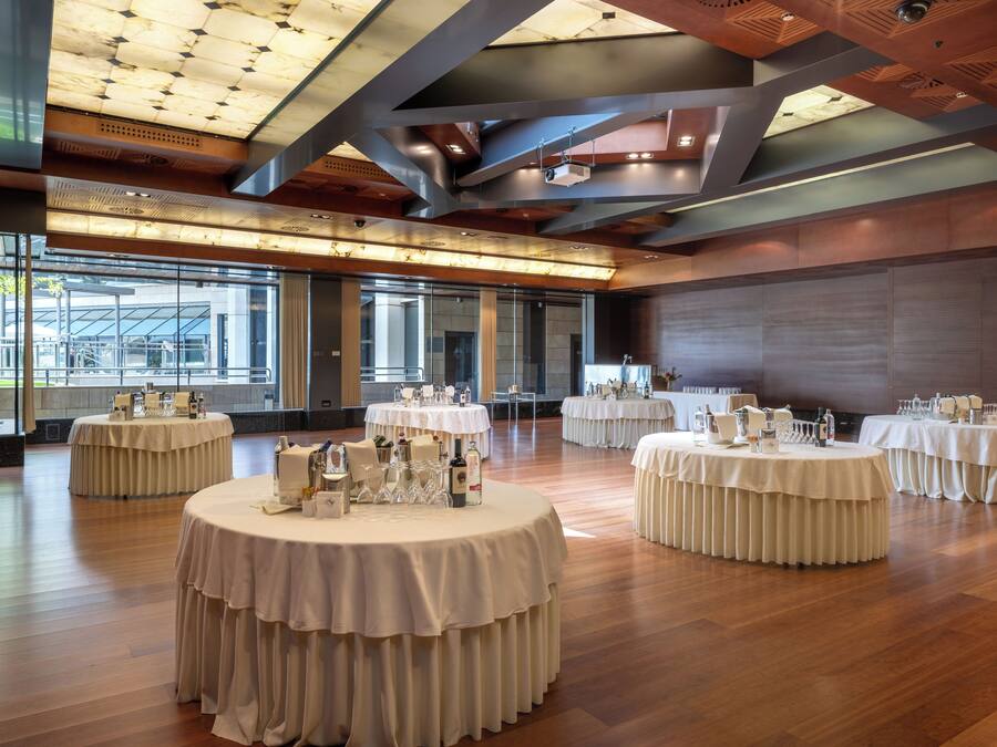 A spacious hall with multiple round tables, each set with white tablecloths and glassware, and a modern ceiling design.
