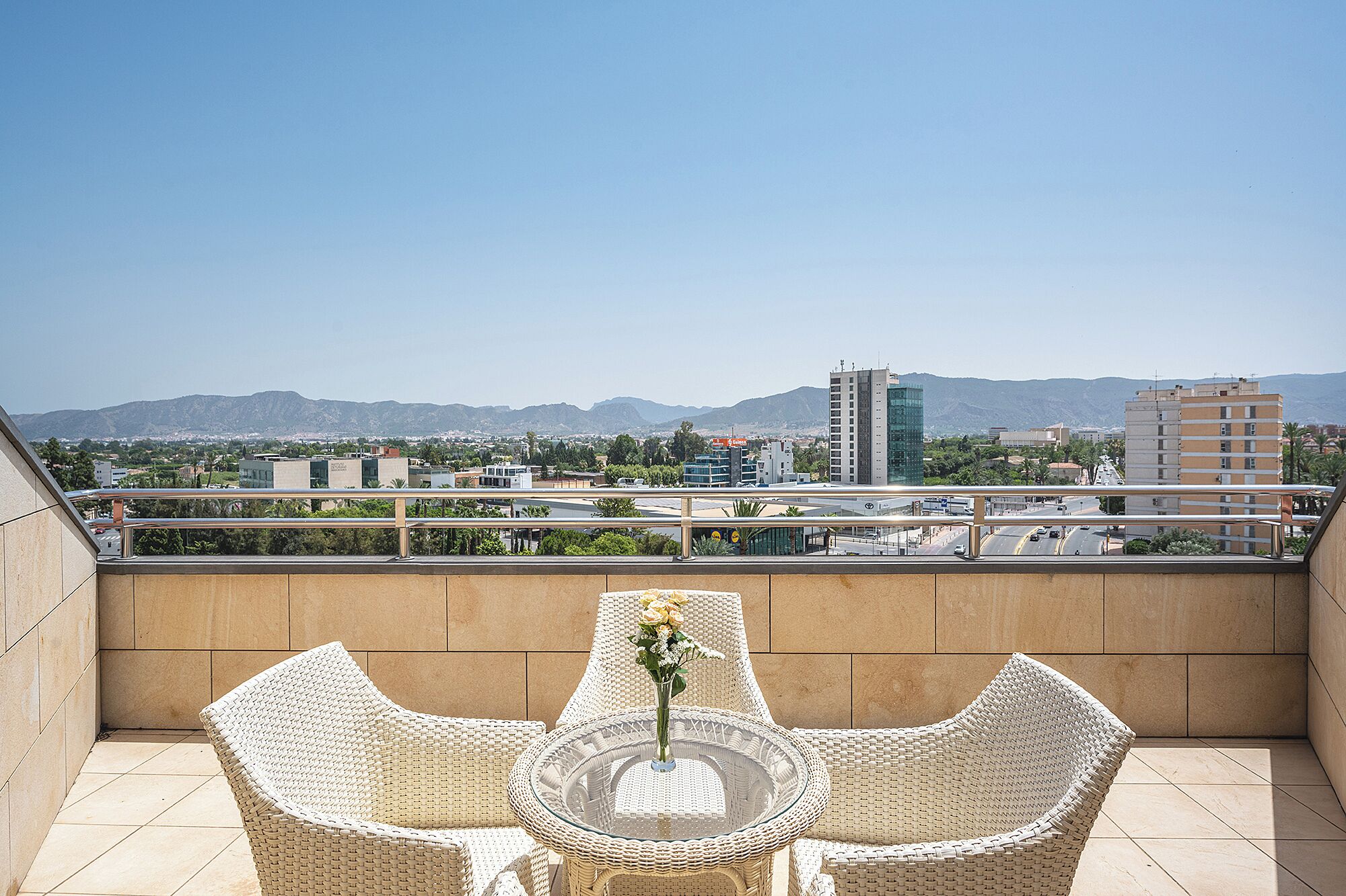 A balcony with wicker furniture and a glass table, overlooking a cityscape with mountains in the distance.