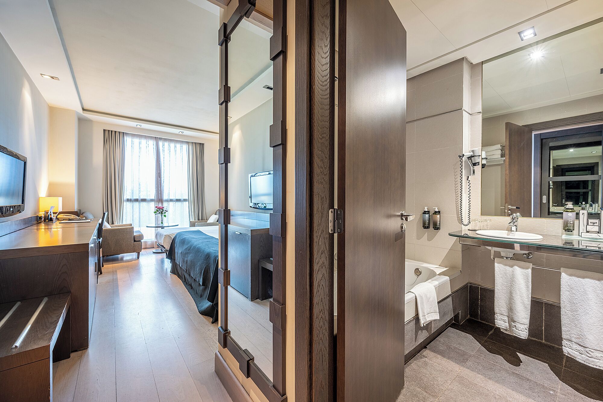 Habitación de hotel con cama, mesita de noche, televisor y baño que incluye espejo, lavabo y toallero.
